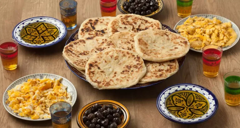 Rustikaler marokkanischer Frühstückstisch mit Spiegeleiern, Oliven, Fladenbrot und süßem Minztee in bunten Gläsern.