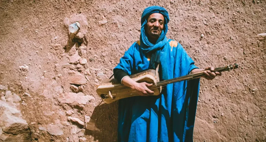 Amazigh musician in blue traditional clothing playing a guembri against an earthen wall in rural Morocco.