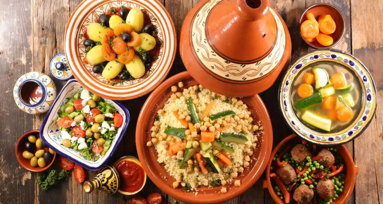 A vibrant Moroccan meal featuring couscous with vegetables, chickpeas, and a variety of fresh salads like tomato and cucumber, served with meatballs, tagine, and olives.