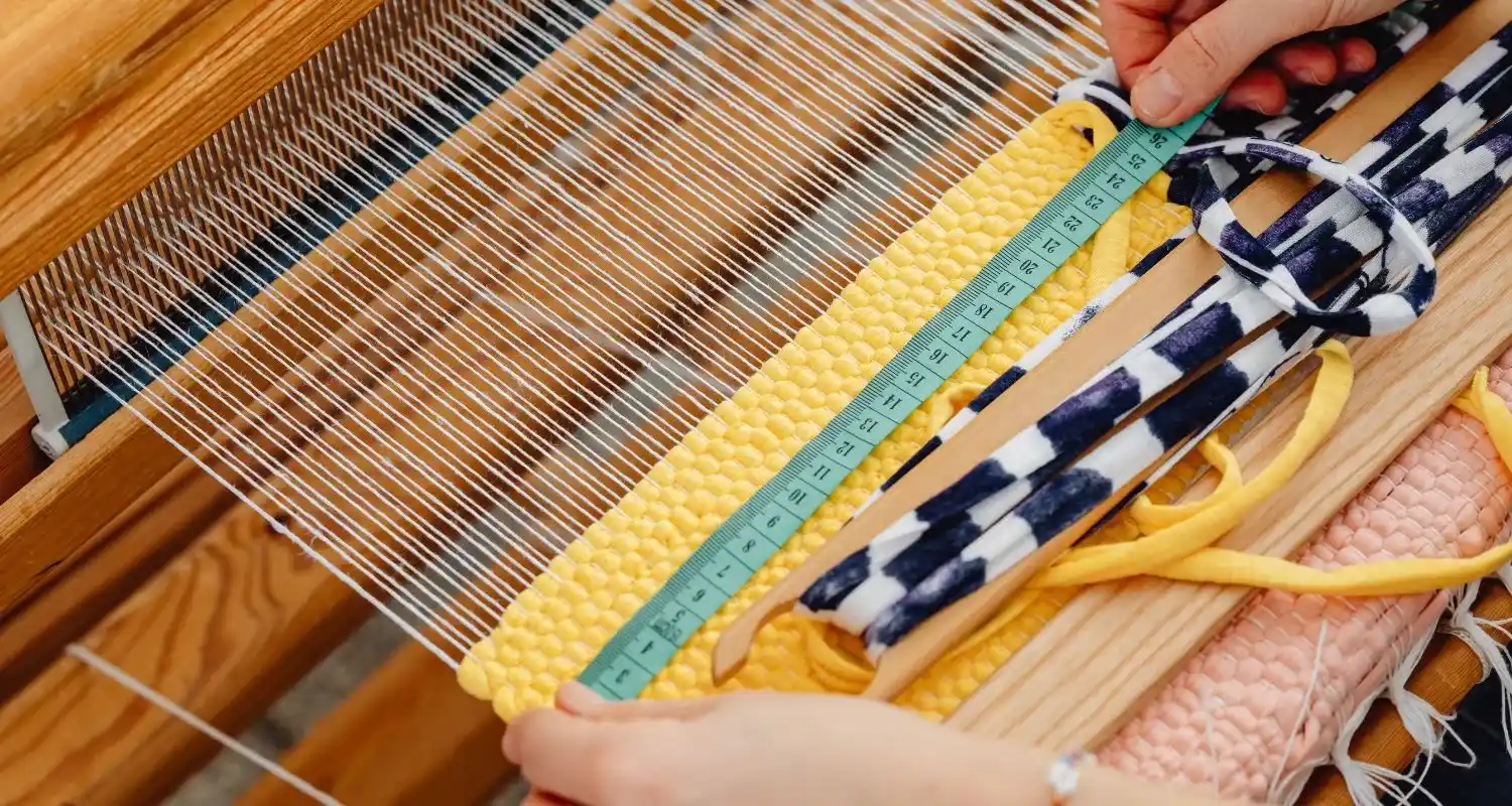 Hands weaving a yellow and blue-and-white textile on a wooden loom with a ruler measuring the weave — the mat-weaving craft of Salé medina