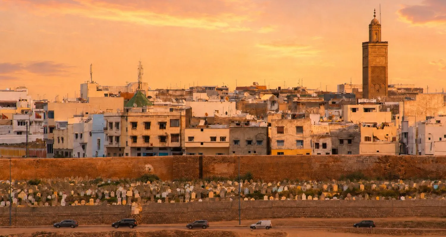 Dense old houses and a minaret of Salé medina rising behind the ramparts at sunset, with a walled cemetery in the foreground